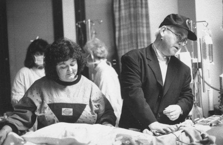 Elton John and Jeanne White stand together beside Ryan White's hospital bed, who was diagnosed with AIDS after a blood transfusion in 1984.