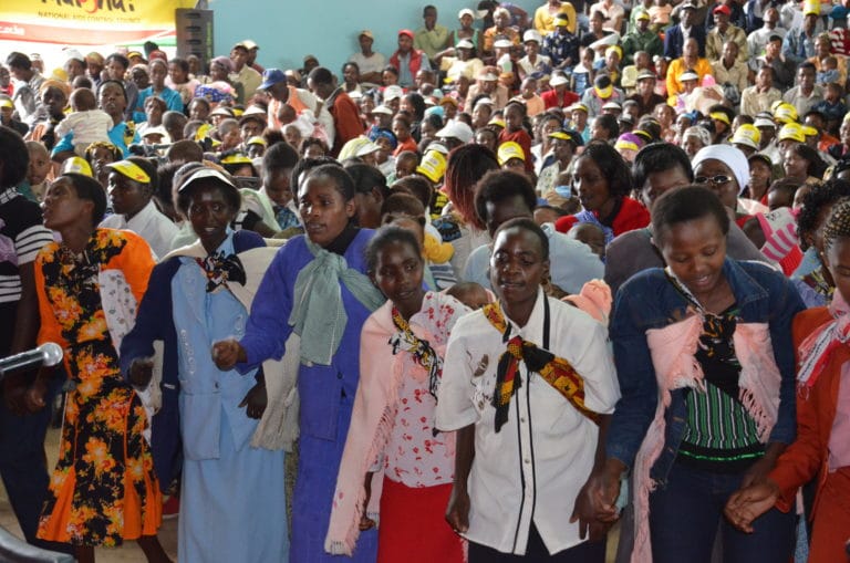 Large gathering with the National AIDS Control Council in Kenya. Front row of community members stand in a line together, holding hands, and singing.