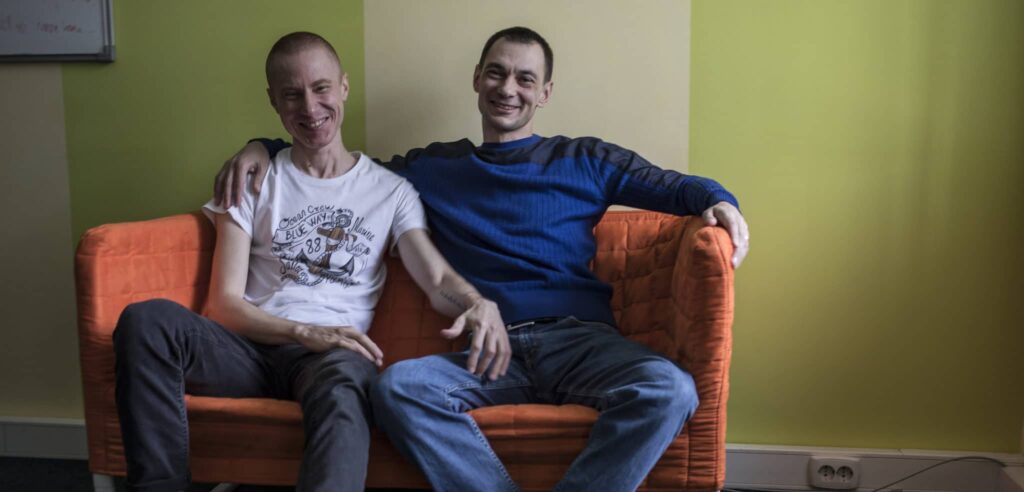 Igor and Sasha sit next to each other in a happy embrace on an orange sofa. They have received support from the Elton John AIDS Foundation's LGBTQ+ work in Eastern Europe and Central Asia.