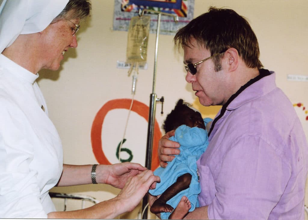Elton John comforts holds and comforts a baby while a nurse attends to them during a visit to a Children's hospital in South Africa.