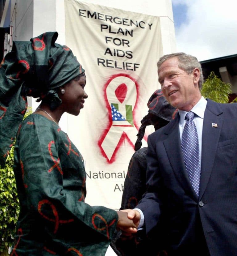 President Bush shakes hands standing in front of a banner that reads 'Emergency Plan for AIDS Relief'.