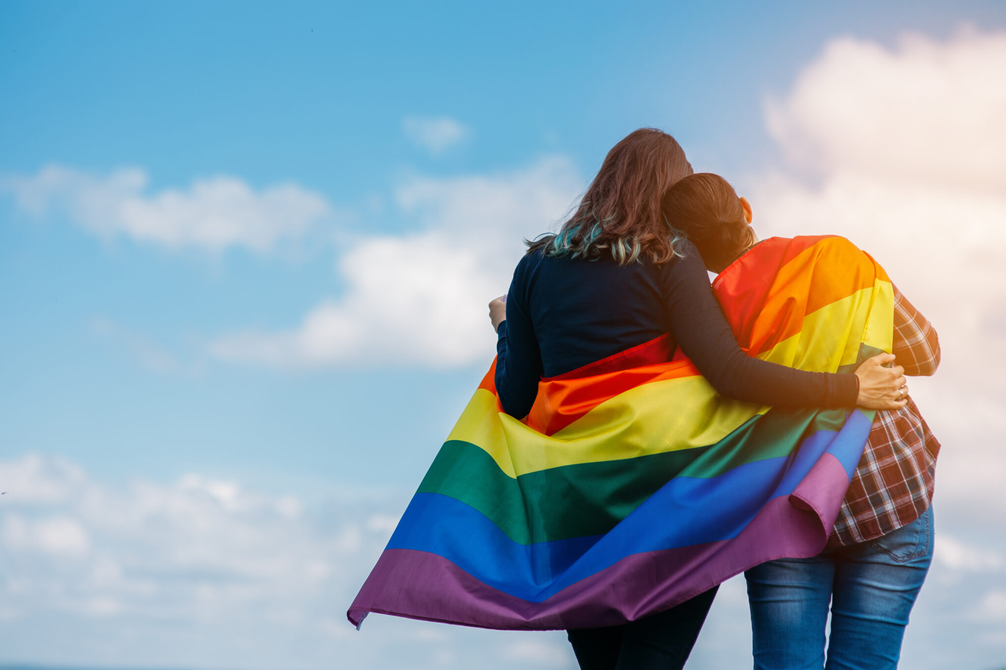 LGBTQ+ couple stand side by side with their backs to the camera with Pride flag draped around them