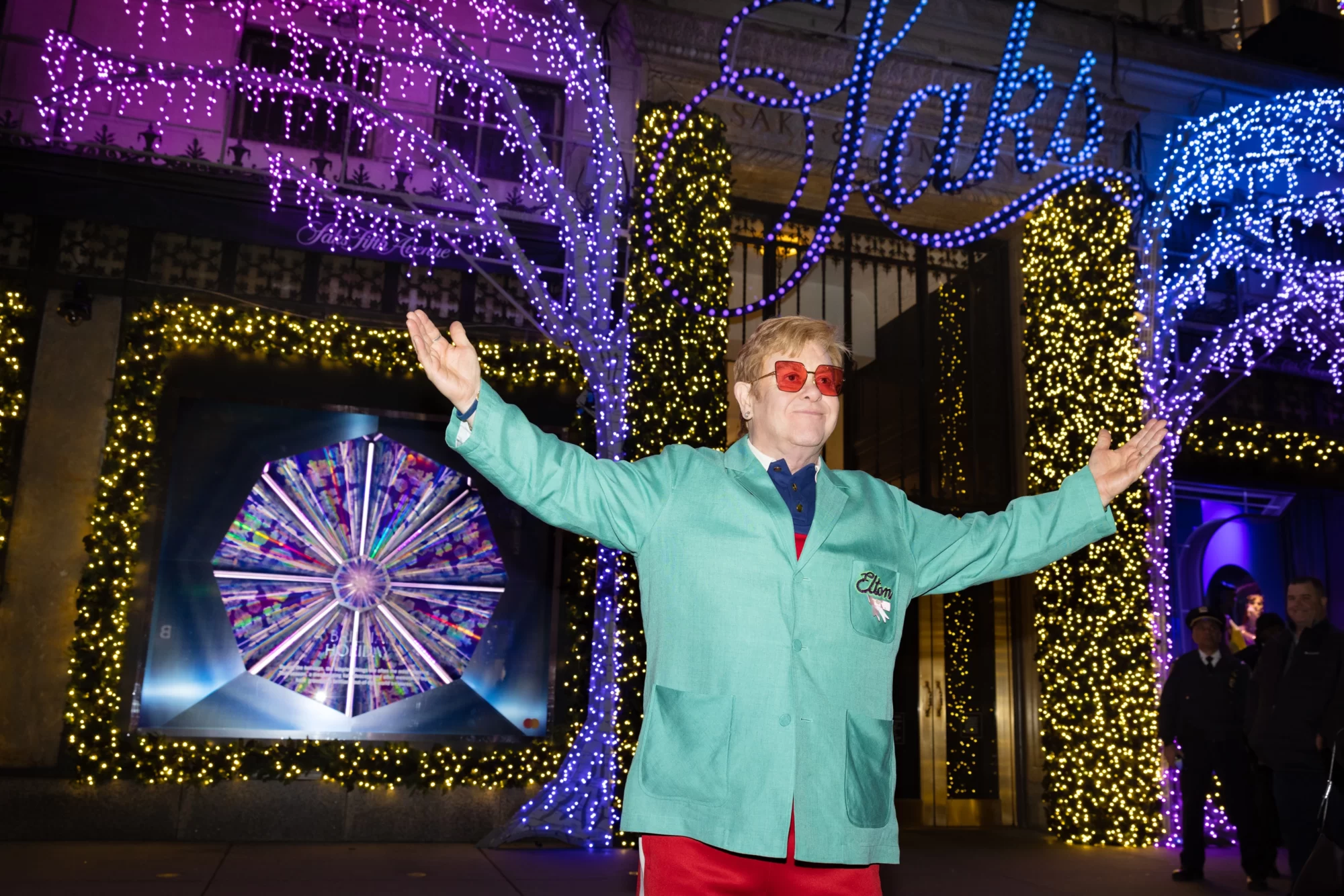 Elton John, wearing a green suit jacket and red trousers, stands in front of a Saks after performing at their holiday window unveiling event in New York.