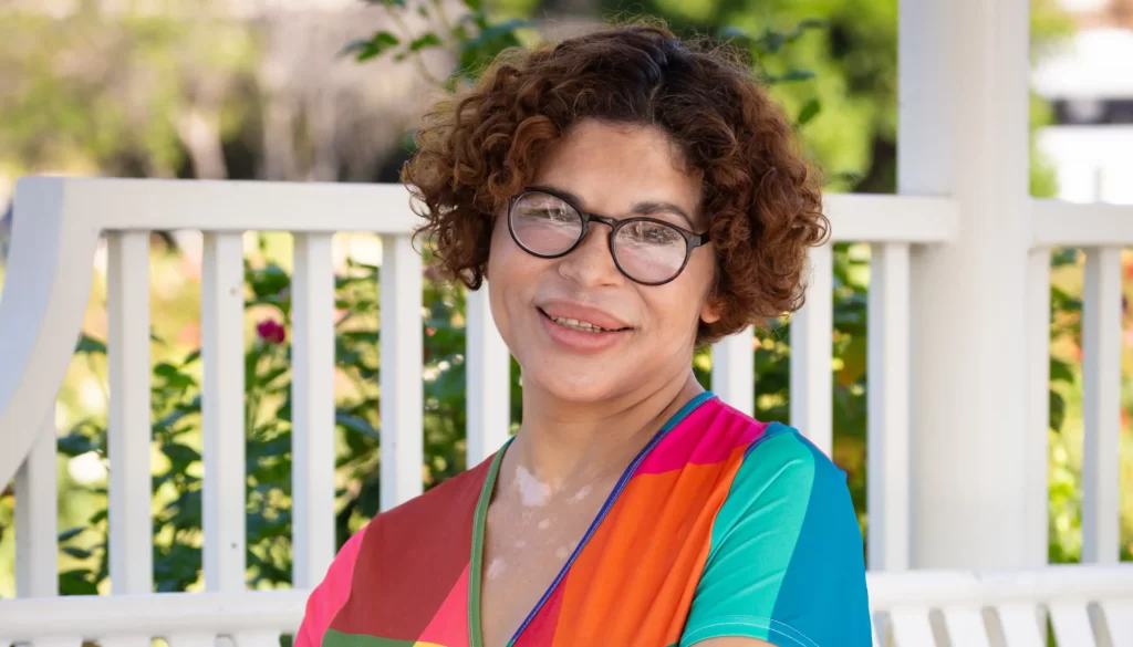 Gina, a migrant transgender woman, poses in a colourful striped dress, glasses and curled hair. She volunteers with our partners to support the LGBTQ+ community.