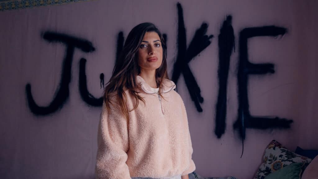 Woman wearing a pink jumper stands in her bedroom with the word 'Junkie' graffitied on the wall behind her.