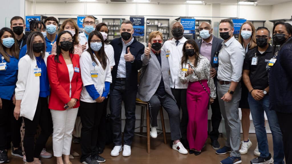 Elton John, David Furnish and Anne Aslett stand with the Walmart Heath team during a visit in Atlanta. All are wearing face masks and Elton and David are giving a thumbs up sign.