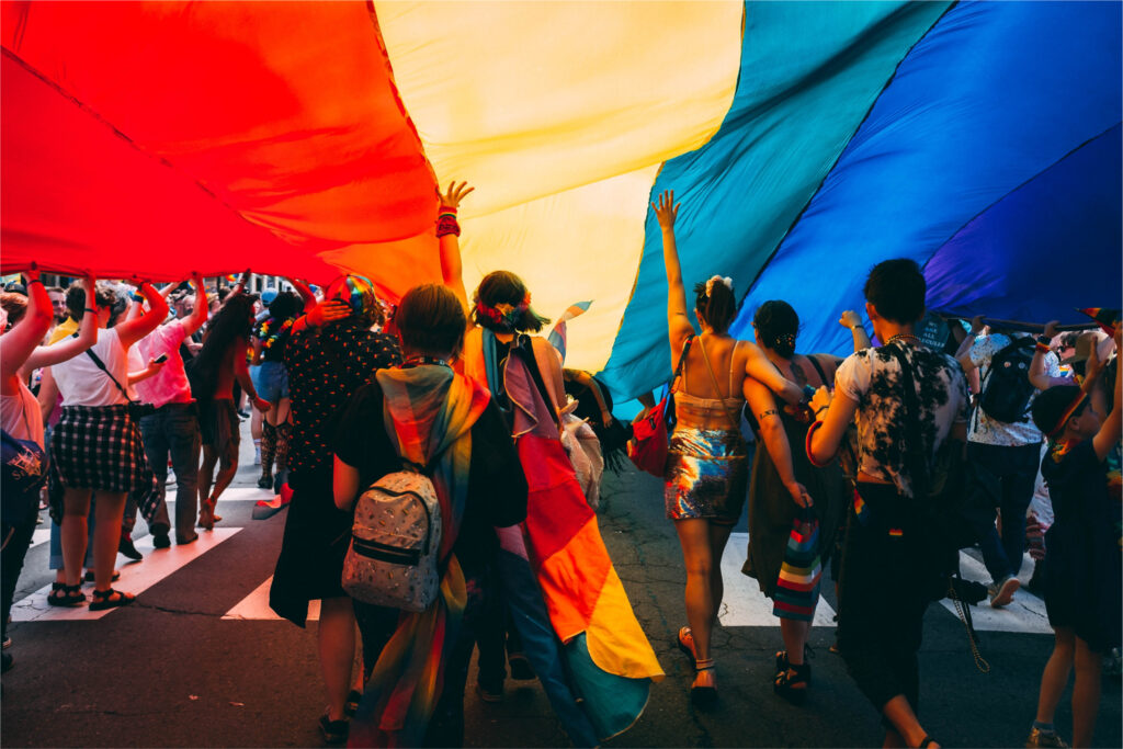 People marching in a Pride parade holding a big Pride flag above them in celebration of the LGBTQ+ community.