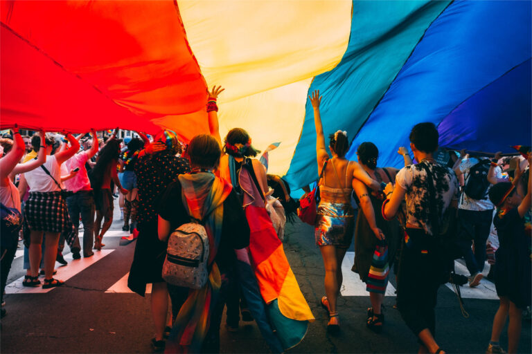 People marching in a Pride parade holding a big Pride flag above them in celebration of the LGBTQ+ community.