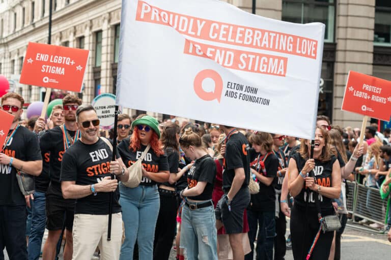 David Furnish and Anne Aslett stand holding a large banner that reads 'Proudly celebrating love to end stigma' with the Elton John AIDS Foundation team in the background