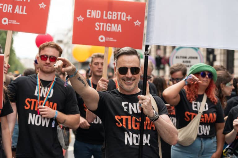 David Furnish points towards the crowd at London Pride, wearing a black top that says 'End Stigma Now'.