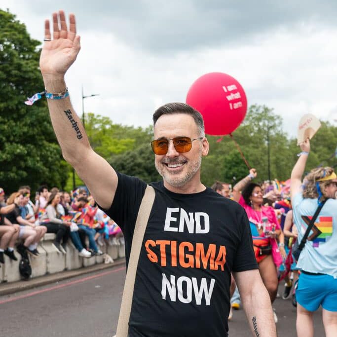 David Furnish waving at the crowd at London Pride, wearing a black top that says 'End Stigma Now'.