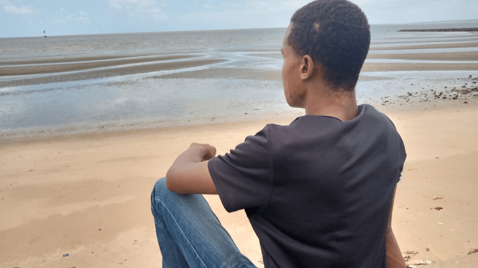Duarte, a young person living with HIV who has been supported through partners of the Elton John AIDS Foundation, sits on a beach in Mozambique looking out at the water.