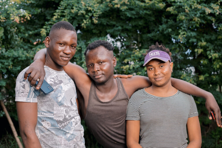Three young men stand together. The person in the middle has his arms around the others.