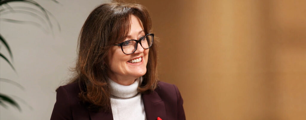 Anne Aslett wears a white jumper and maroon blazer, smiles at interviewer who is off screen.