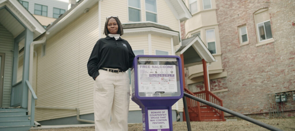 Woman stands next to a free naloxone dispenser.
