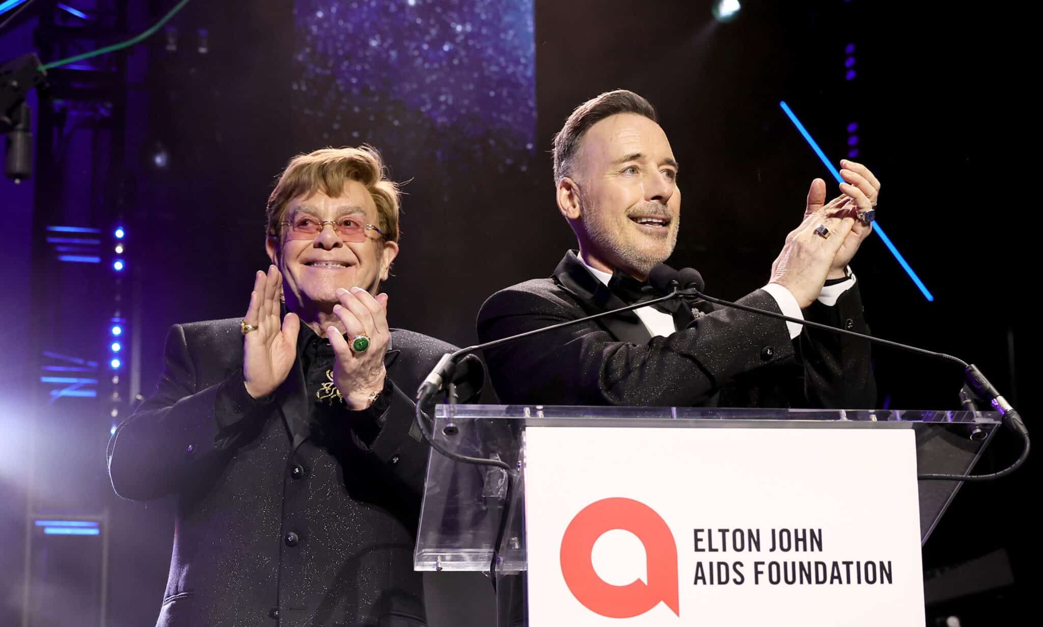 Elton John and David Furnish smile and clap standing behind Elton John AIDS Foundation podium at the Academy Awards Viewing Party.