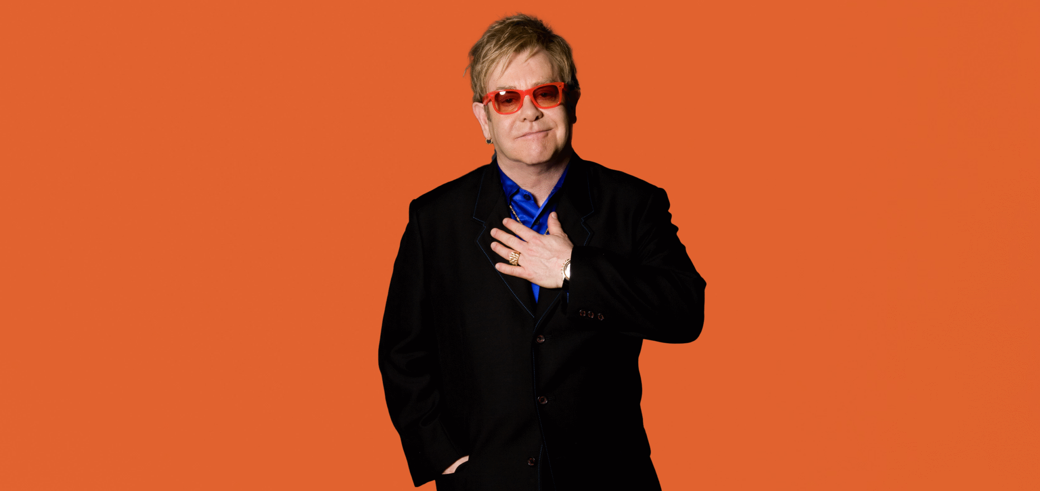 Elton John wearing orange glasses and a black suit with his hand over his heart.