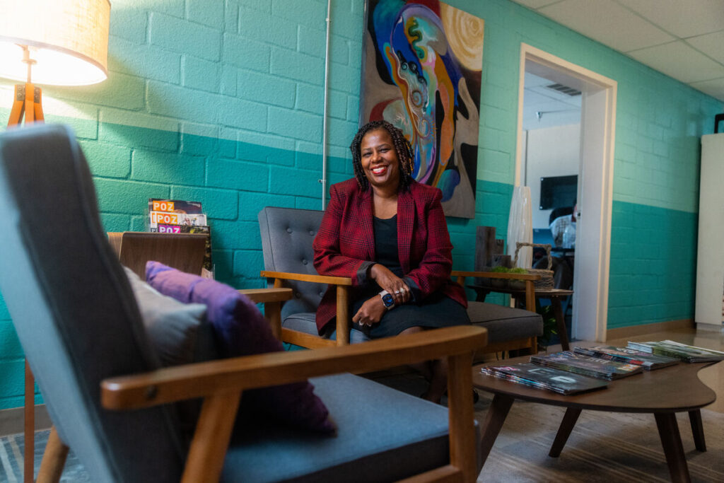 Woman wearing a red blazer poses for a photo seated, and smiling against a turquoise wall.
