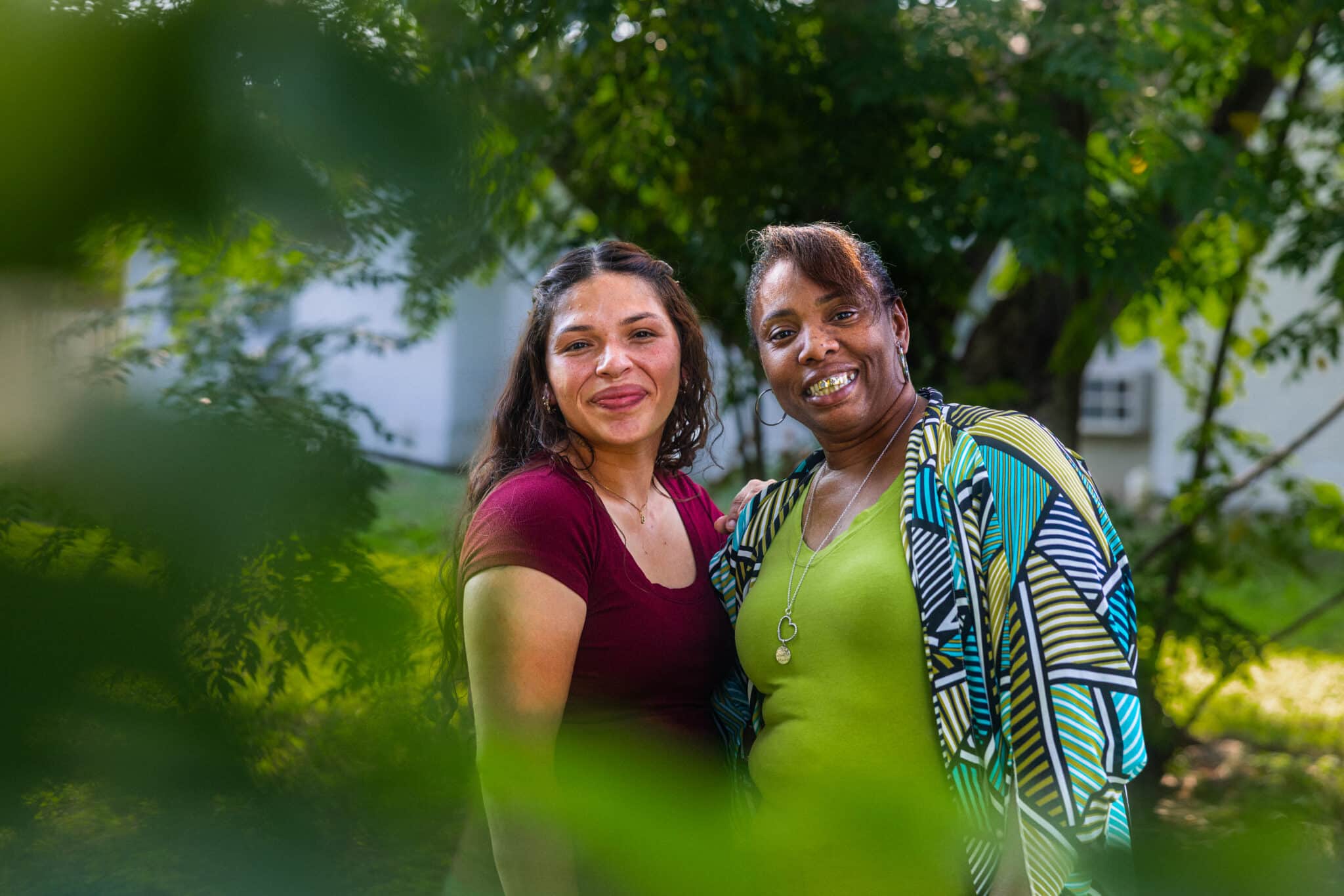 Ashley, wearing a burgundy top, and Violeta, wearing a green patterned top, stand side by side smiling towards the camera. They are in the garden surrounded by greenery.