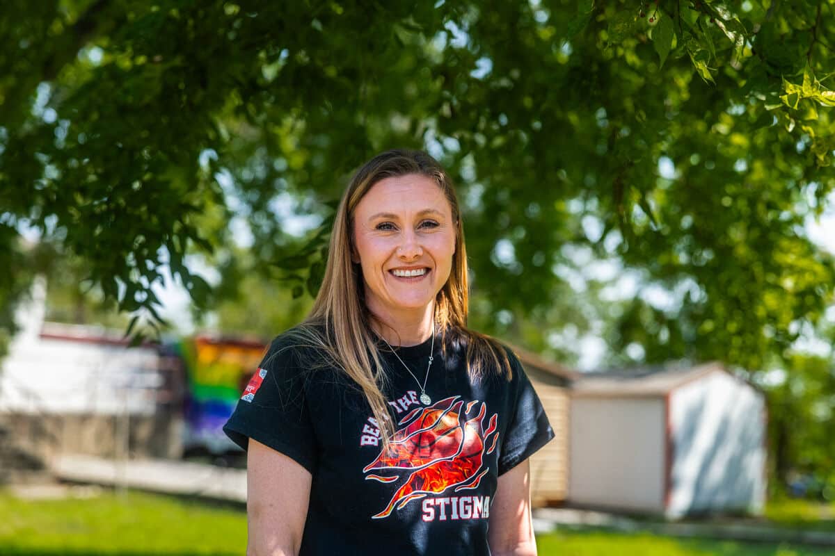 A woman smiles wearing a black t-shirt. She stands underneath a tree.