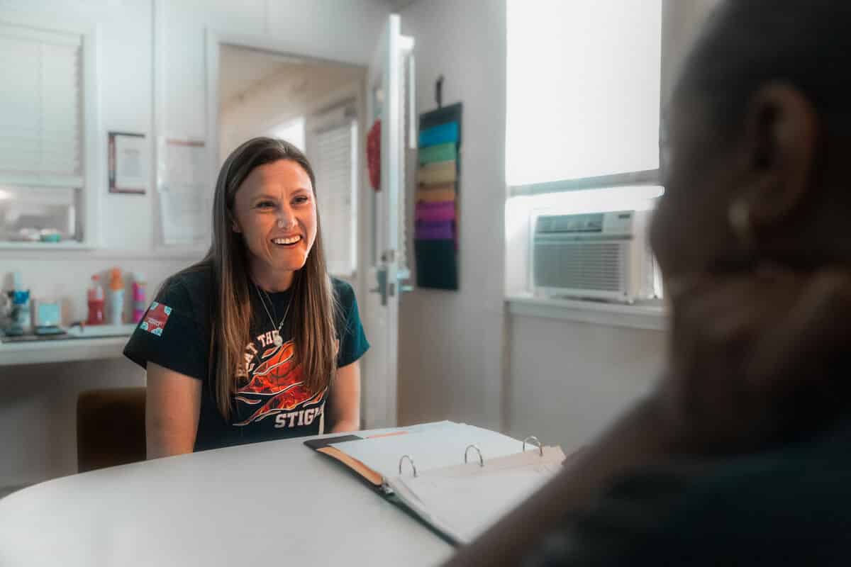A woman smiles whilst talking to someone whose back we see. There is a table between them and they are in an office environment.