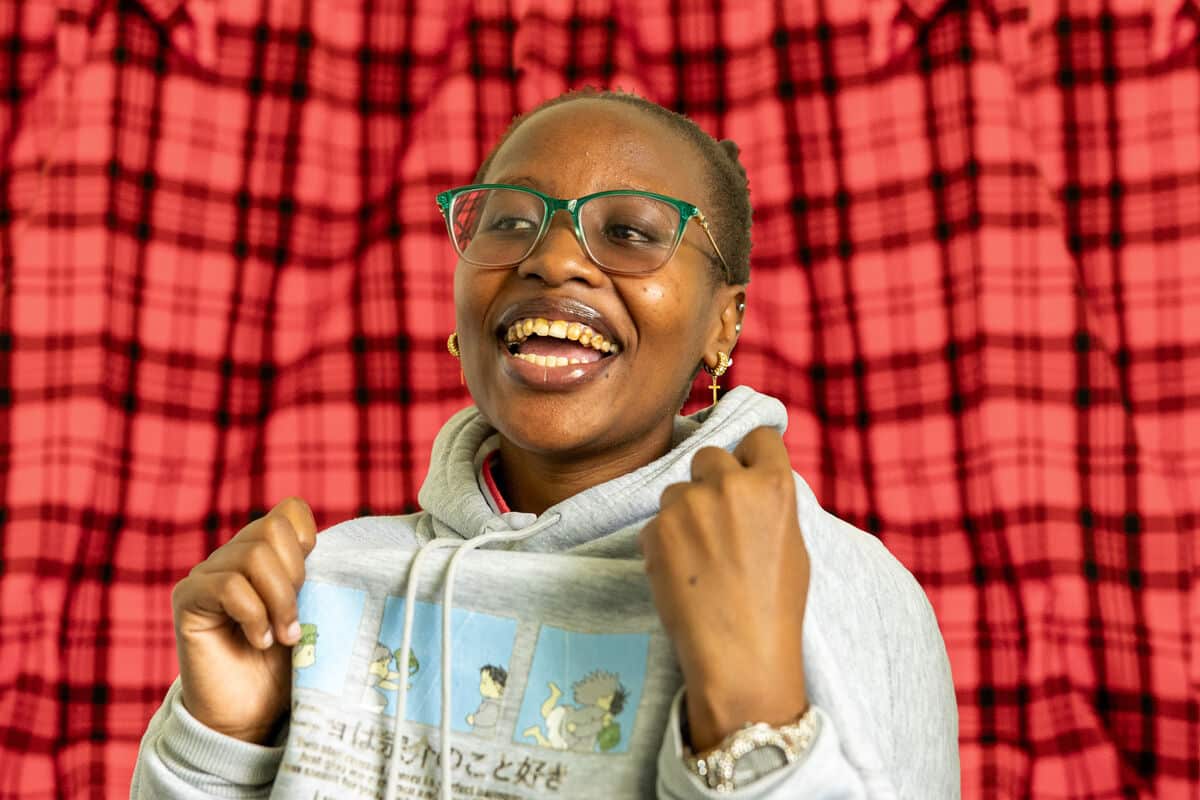 A joyful person wearing glasses and a hoodie smiles widely with clenched fists raised in excitement, standing in front of a red checkered curtain background.