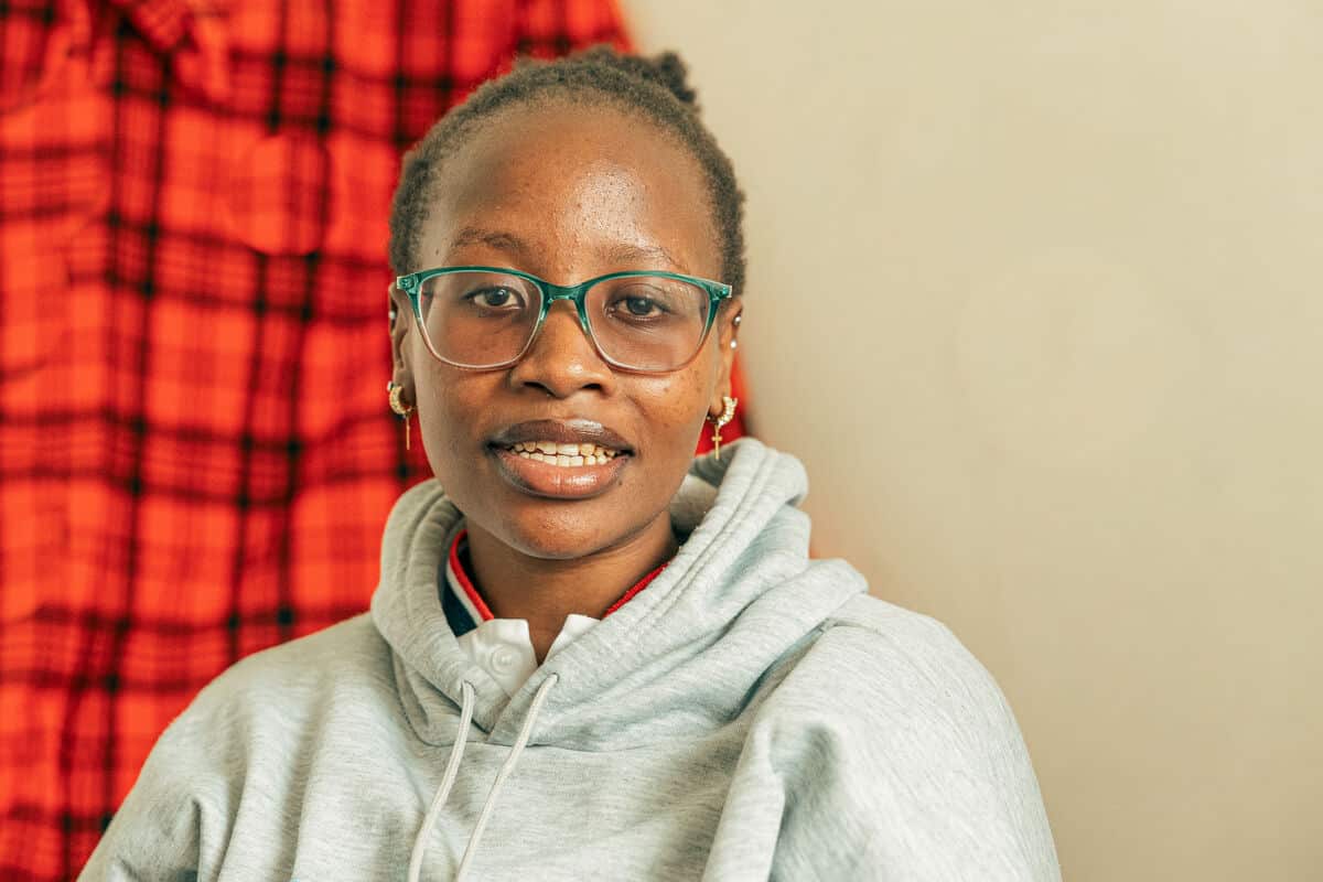 A young woman wearing glasses and a gray hoodie, smiling gently while sitting indoors against a plain beige wall with a red patterned fabric in the background.